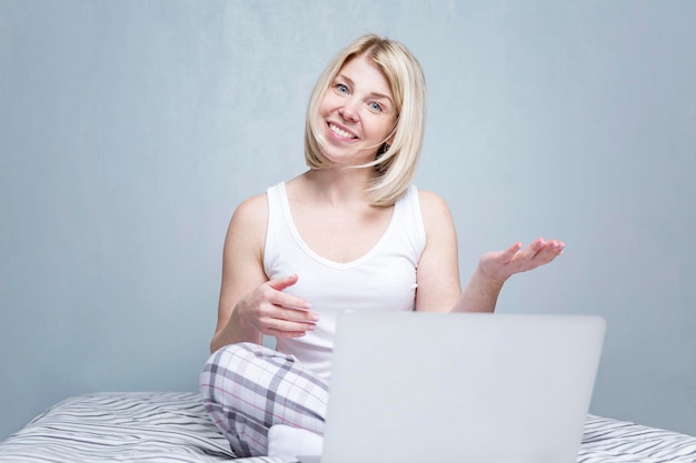 A young woman sits on a bed with a laptop and chats online Beautiful blonde in pajamas against a gray wall Remote work and communication in social networks