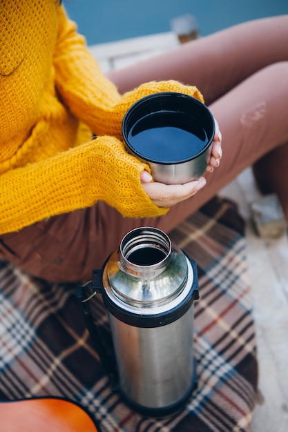 Young woman sits on a bridge on a lake with an autumn landscape and drinks hot tea from a thermos. Toning.