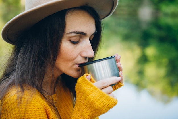 Young woman sits on a bridge on a lake with an autumn landscape and drinks hot tea from a thermos. Toning.