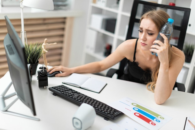 A young woman sits at a computer desk in the office and puts a bottle of water to her head.