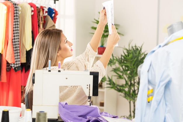 Young woman sits in front of the sewing machine and looking at sketches of clothing. In front of her is a mannequin, and behind are pieces of clothing arranged on a hanger.