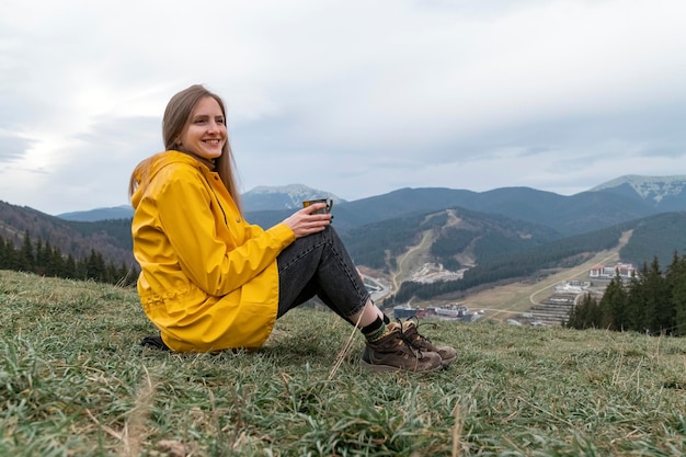 Young woman sits on hillside drinking tea and smiling Portrait of tourist girl in yellow jacket on the mountains