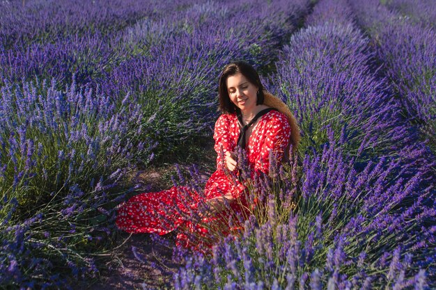 A young woman sits in a lavender field with a straw hat hanging around her neck