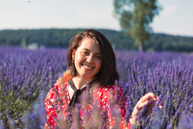 A young woman sits in a lavender field with a straw hat hanging around her neck