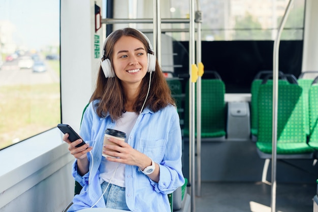 Young woman sits in the modern city bus, listens to music, drinks coffee and looks out from the window.