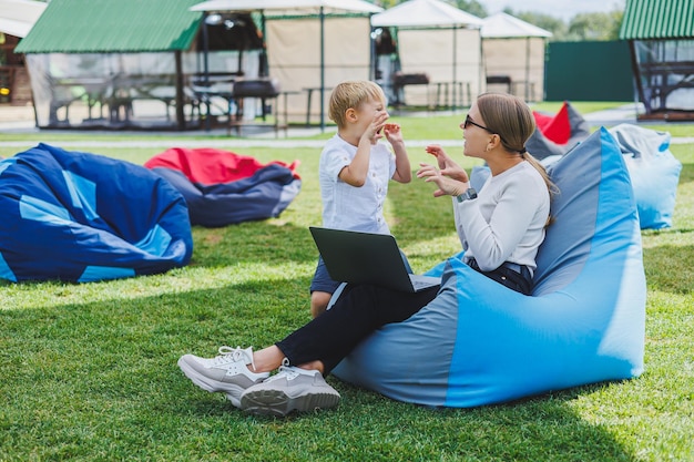 A young woman sits on a soft chair in the park and works on a laptop while her son plays next to her Working outdoors on maternity leave