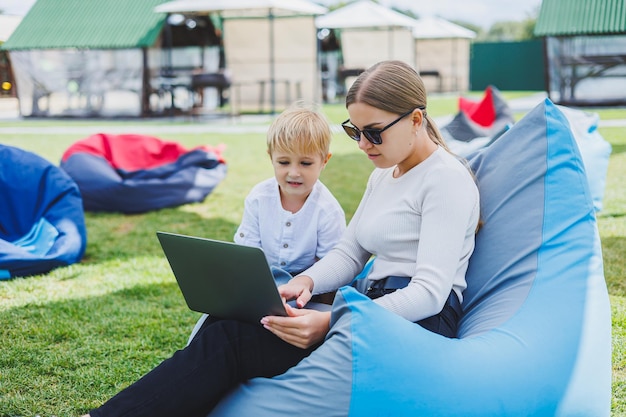 A young woman sits on a soft chair in the park and works on a laptop while her son plays next to her Working outdoors on maternity leave