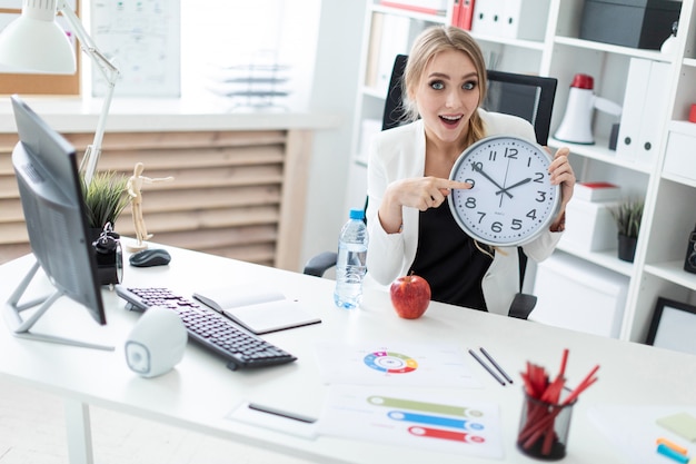 A young woman sits at a table in the office and points a finger at the clock face.