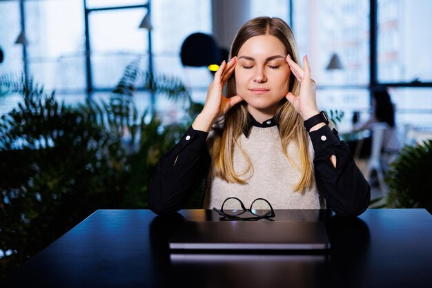 A young woman sits at a table with a closed laptop and massages her head from hard work and headaches Woman suffering from migraines