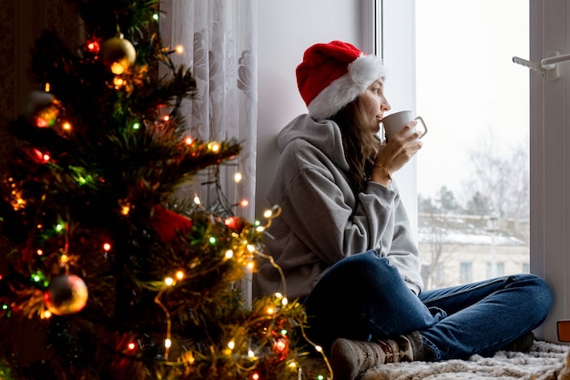 A young woman sits at the window in a decorated house for christmas, looks out the window
