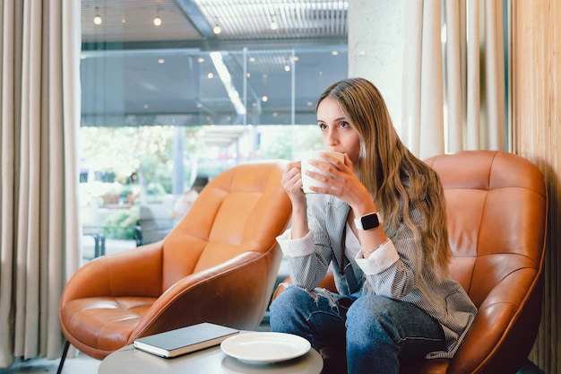 Young woman sitting on an armchair, drinking coffee in an open workplace, waiting for a late friend