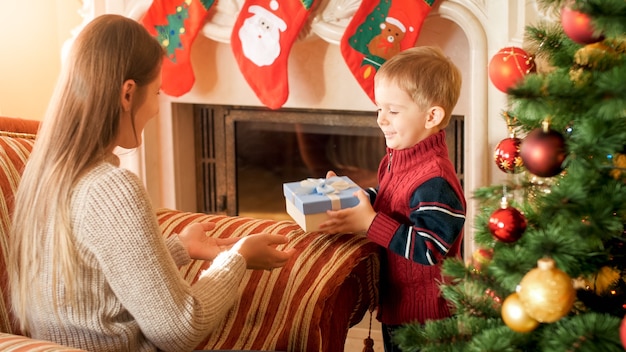 Young woman sitting in armchair in living room and giving box with Christmas present to her little son