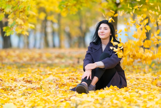 Young woman sitting in an autumn park amongst colorful yellow leaves smiling as she daydreams