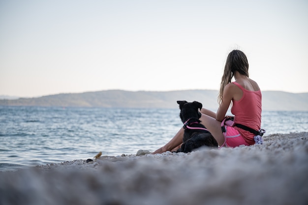 Young woman sitting on beautiful pebble beach with her black shepherd dog lying next to her.