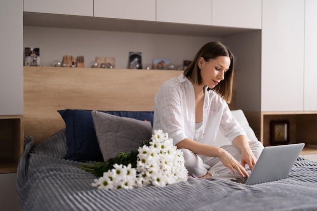 Young woman sitting on the bed wearing pajamas with pleasure enjoying white flowers chatting using laptop