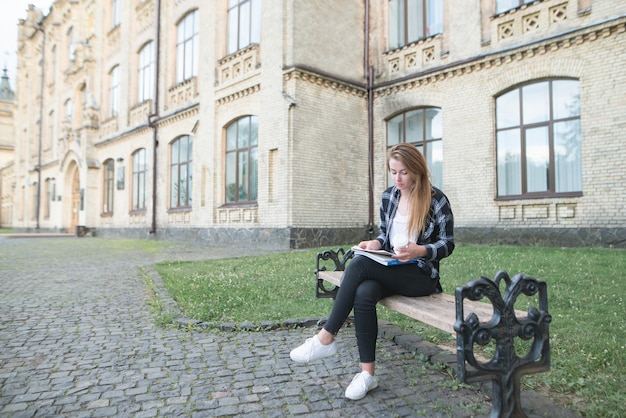 Photo young woman sitting on a bench on the background of a university building with a book in her hands and studying