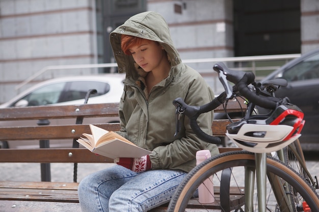 Young woman sitting on a bench near her bicycle, reading a book