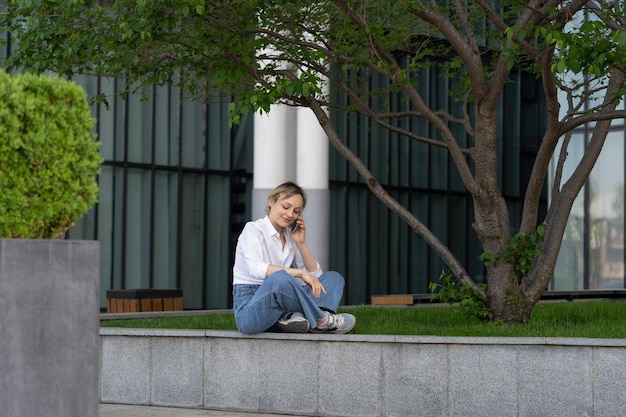 Young woman sitting on bench in spring park under tree outdoors resting talking by mobile cell phone