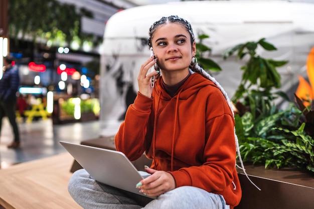 A young woman sitting on bench with her laptop talking with her phone in a shopping mall