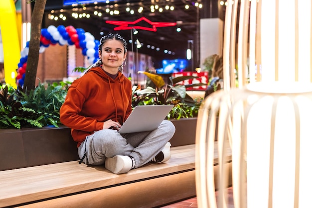 A young woman sitting on bench with her legs crossed using her laptop with a lamp on front in a shopping mall