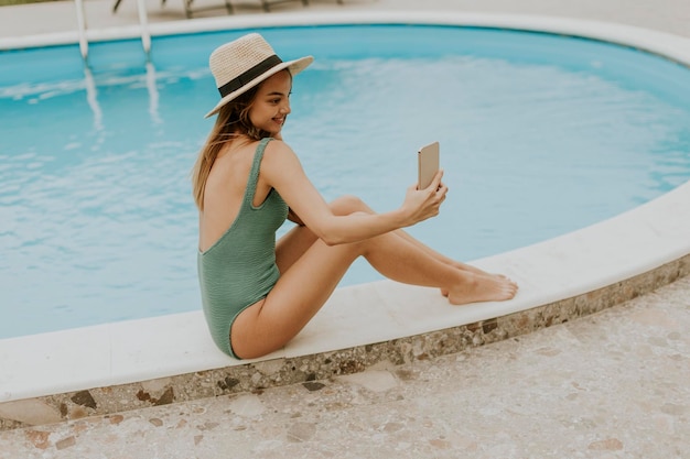 Young woman sitting by the swimming pool and taking selfie photo with mobile phone in the house backyard