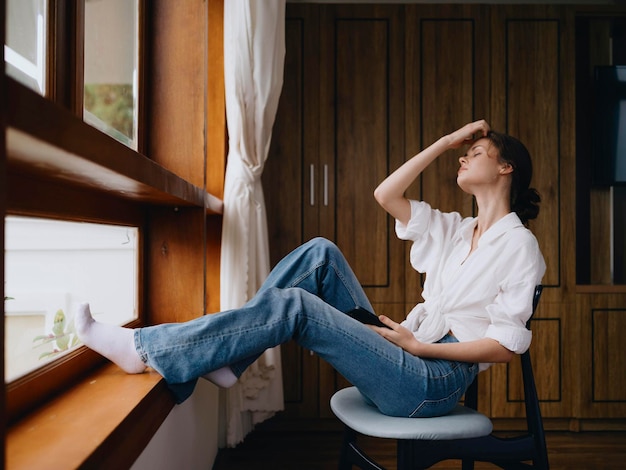 Young woman sitting by the window with phone in hand smile looking online spring mood home simple clothes home cozy atmosphere aesthetic lifestyle