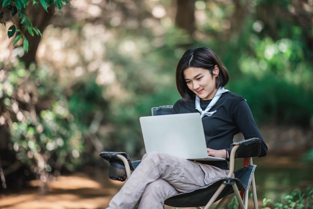 Young woman sitting on camping chair and use laptop computer while relax on camping in forest copy space