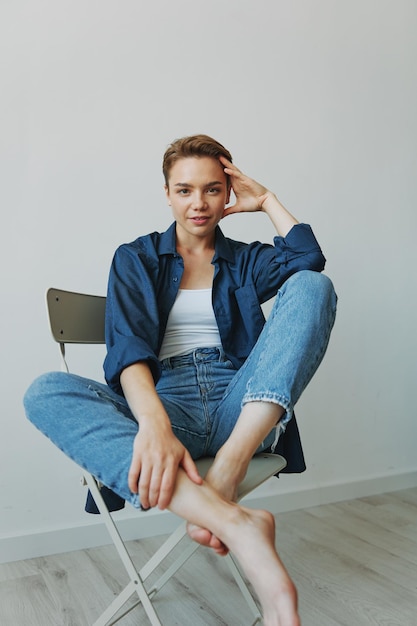 A young woman sitting in a chair at home smiling with teeth with a short haircut in jeans and a denim shirt on a white background Girl natural poses with no filters