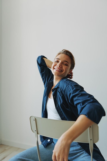 A young woman sitting in a chair at home smiling with teeth with a short haircut in jeans and a denim shirt on a white background Girl natural poses with no filters