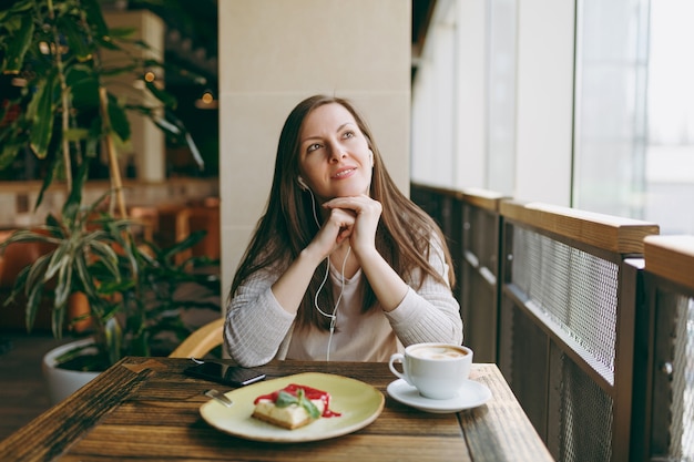 Young woman sitting in coffee shop at table with cup of cappuccino, cake, relax in restaurant during free time. Young female listen music in earphones on mobile phone, rest in cafe. Lifestyle concept.