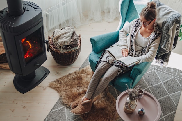 Young woman sitting in a cozy armchair with a warm blanket reading a book