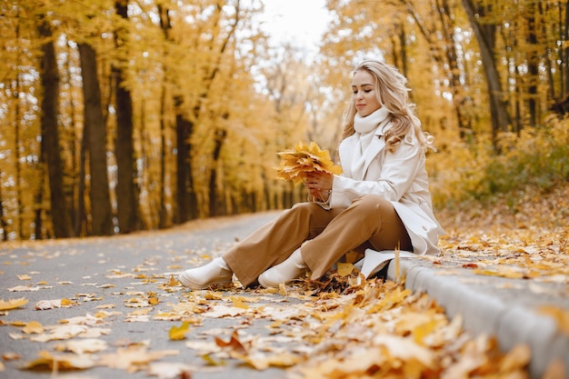 Young woman sitting on a curb in autumn forest. Blonde woman holding a yellow leaves. Girl wearing beige coat and brown trousers.