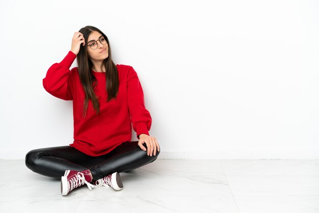 Young woman sitting on the floor isolated on white background having doubts and with confuse face expression