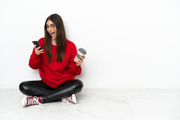 Young woman sitting on the floor isolated on white background holding coffee to take away and a mobile