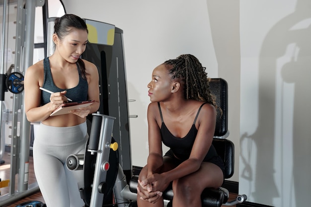 Young woman sitting on gym machine and listening to recommendations of her fitness trainer