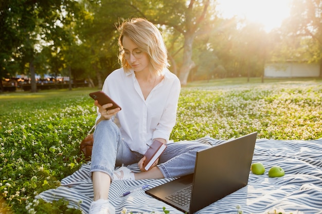 Young woman sitting on the lawn in the park uses laptop and smartphone