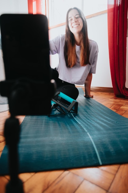 Young woman sitting on mat in sportswear starting recording herself with a mobile phone on a tripod while streaming to all of her followersvideo via smartphone for her vlog