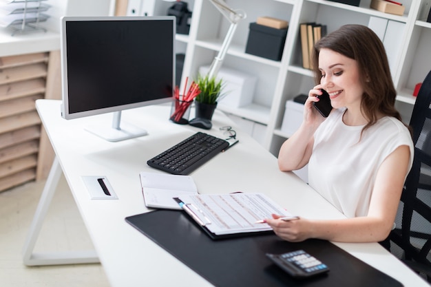 A young woman sitting in the office at the computer Desk, talking on the phone and making notes on the sheet.