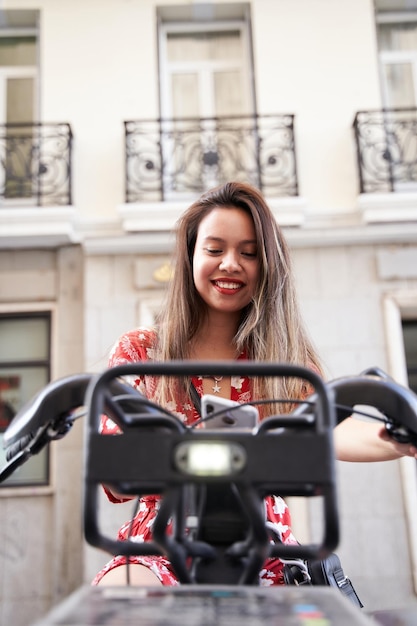 Young woman sitting on a rental bike smiling with her cell phone