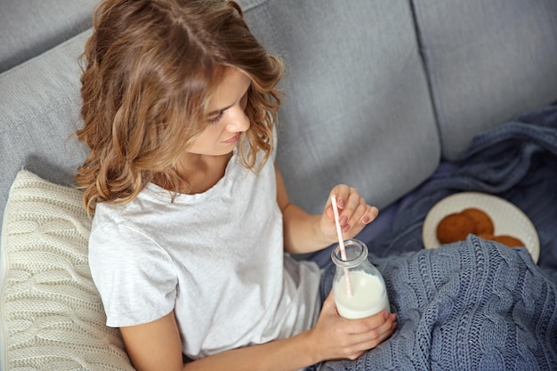 Young woman sitting on sofa with bottle of milk and cookies