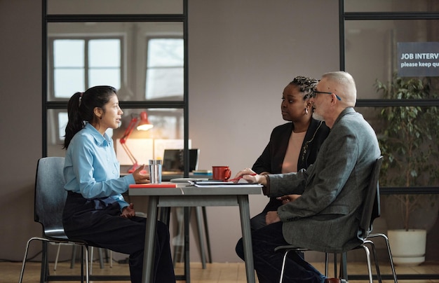 Young woman sitting at table with employers and presenting her resume during job interview at office