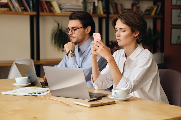 Young woman sitting at the table with laptop and cup of coffee thoughtfully using cellphone Business colleagues working in modern office