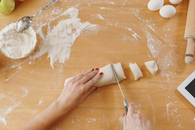 A young woman sitting at a table with tablet, cuts a dough with a knife into pieces at home in the kitchen. Cooking home. Prepare food. Top view.