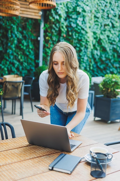 Young woman sitting on the terrace of a cafe on the street of a modern cafe Woman sitting at table with phone and using laptop outdoors Remote work in a summer cafe