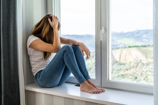 Young woman sitting in the window of her house looks out. Concept of staying at home.