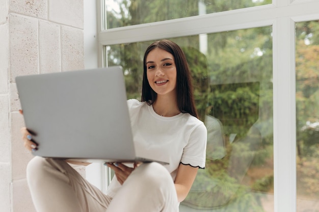 Young woman sitting on the windowsill and working online using her laptop in the room