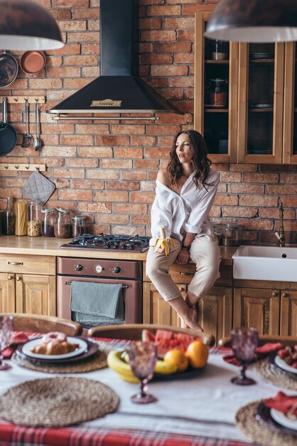 Young woman sitting with a banana in the kitchen on the table.