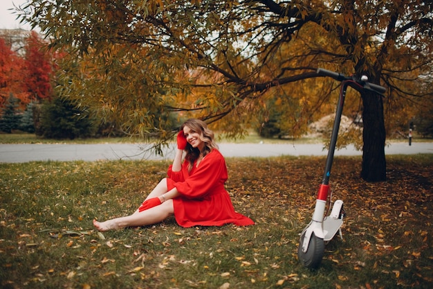 Young woman sitting with electric scooter in red dress at the autumn city park