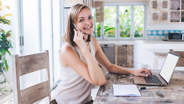 young woman sitting at wooden table with laptop on it at Bali Villa and smiling talking on phone