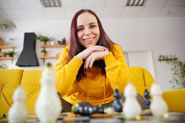 Young woman sitting on yellow sofa and playing chess in room Female playing in logical board game with herself